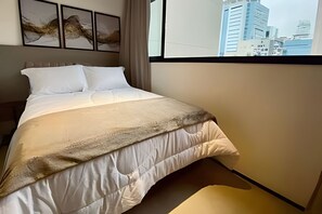 Una cama con edredón blanco y una manta manchada, cabecero y ventana con vista a la ciudad.