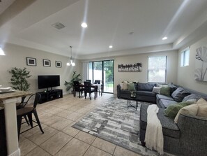 A modern living room with a flat-screen TV, a dining area with a table and chairs, a gray sofa with cushions, and a patterned rug. - Modern Champions Gate Retreat - 2 Primary Suites - 5 Beds - Sleeps 10 (Davenport)