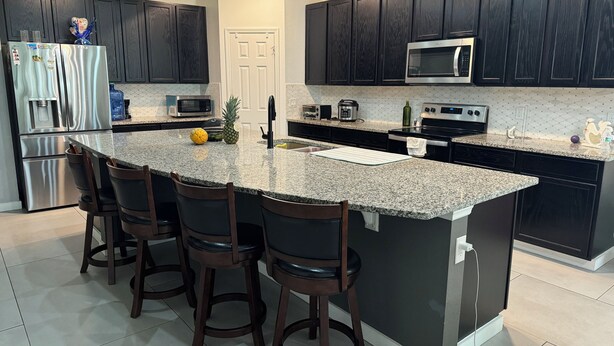 A modern kitchen with dark cabinetry, stainless steel appliances, a granite countertop, and bar stools. - House for rent, 12 minutes from Orlando Airport and 25 minutes from the parks. (Orlando)