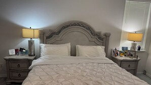 A neatly made bed with a tufted white duvet, two bedside tables with lamps, a decorative headboard, and a window with blinds. - House for rent, 12 minutes from Orlando Airport and 25 minutes from the parks. (Orlando)