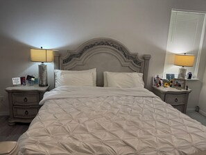 Ein ordentlich bezogenes Bett mit einer gesteppten weißen Decke, zwei Nachttische mit Lampen, ein dekorativer Kopfteil und ein Fenster mit Jalousien.