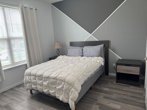 Un dormitorio con cama, mesita de noche, lámpara y ventana con cortinas.