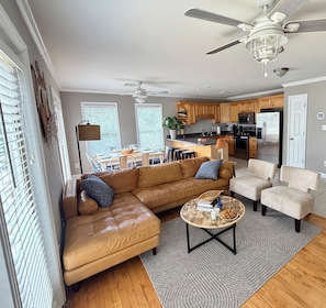 A modern living room with a leather sofa, a coffee table, and a dining area with a kitchen in the background. - Where Isle Be•Relax & Unwind• 4 bed• 4 bath• Sleeps 8 (Emerald Isle)