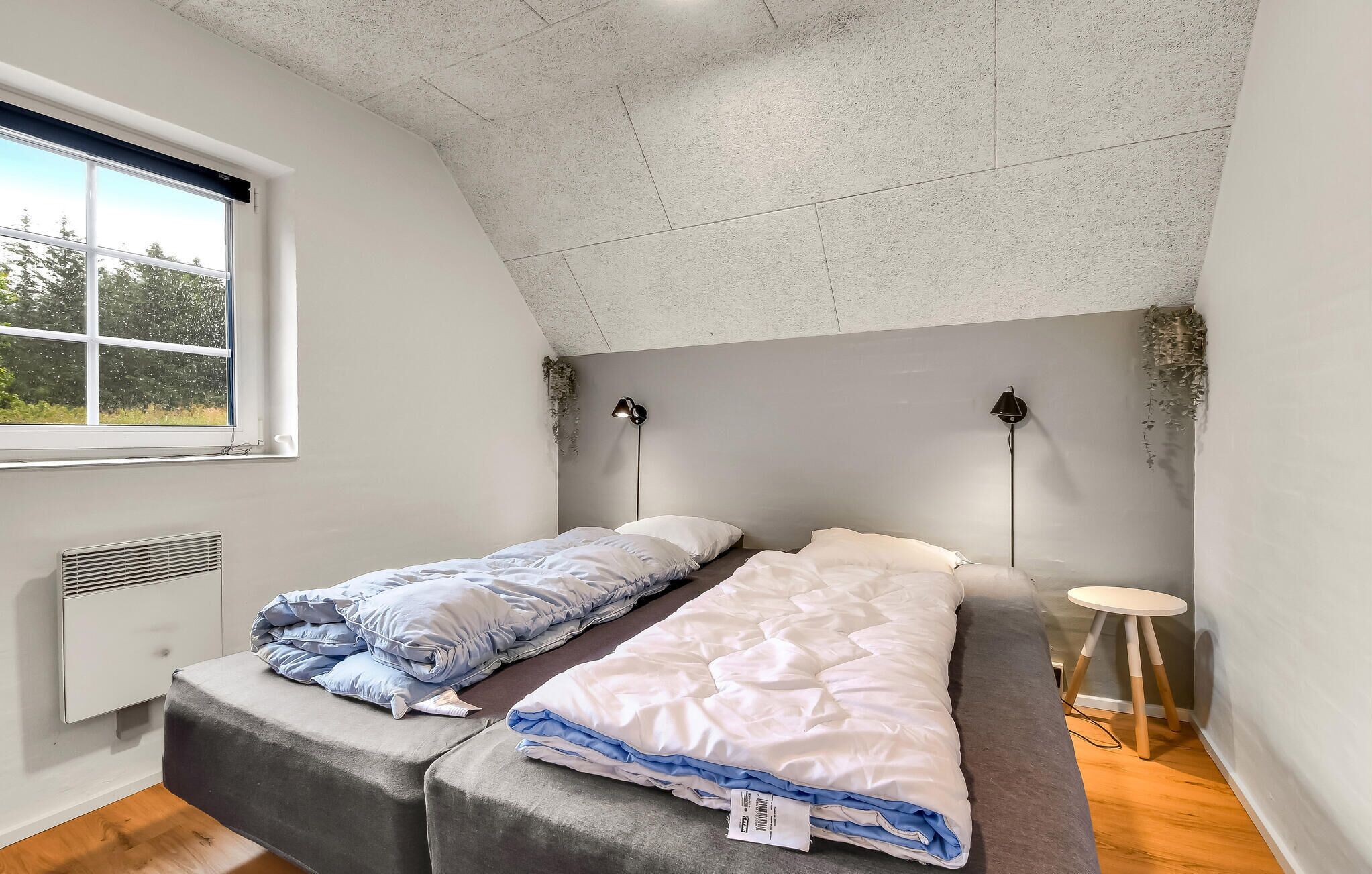 Et værelse med skråvæg, et vindue der ud til grønne omgivelser, en seng med hvid madras og blåt sengetøj, et natbord og en lampe monteret på væggen.