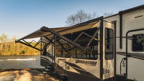 A recreational vehicle with a large awning, a ramp, and a water body in the background. - The Coosa Nova at River Rocks Landing (Gadsden)