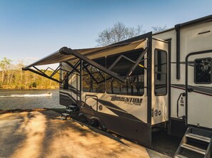 Ein Wohnmobil mit großem Markisenvorzelt, einer Auffahrtrampe und einem See im Hintergrund.