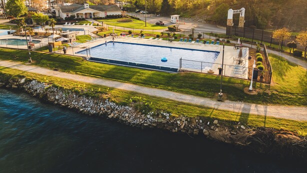 A large rectangular swimming pool with a blue inflatable float, surrounded by a grassy area with lounge chairs and umbrellas, and a small building in the background. - The Coosa Nova at River Rocks Landing (Gadsden)