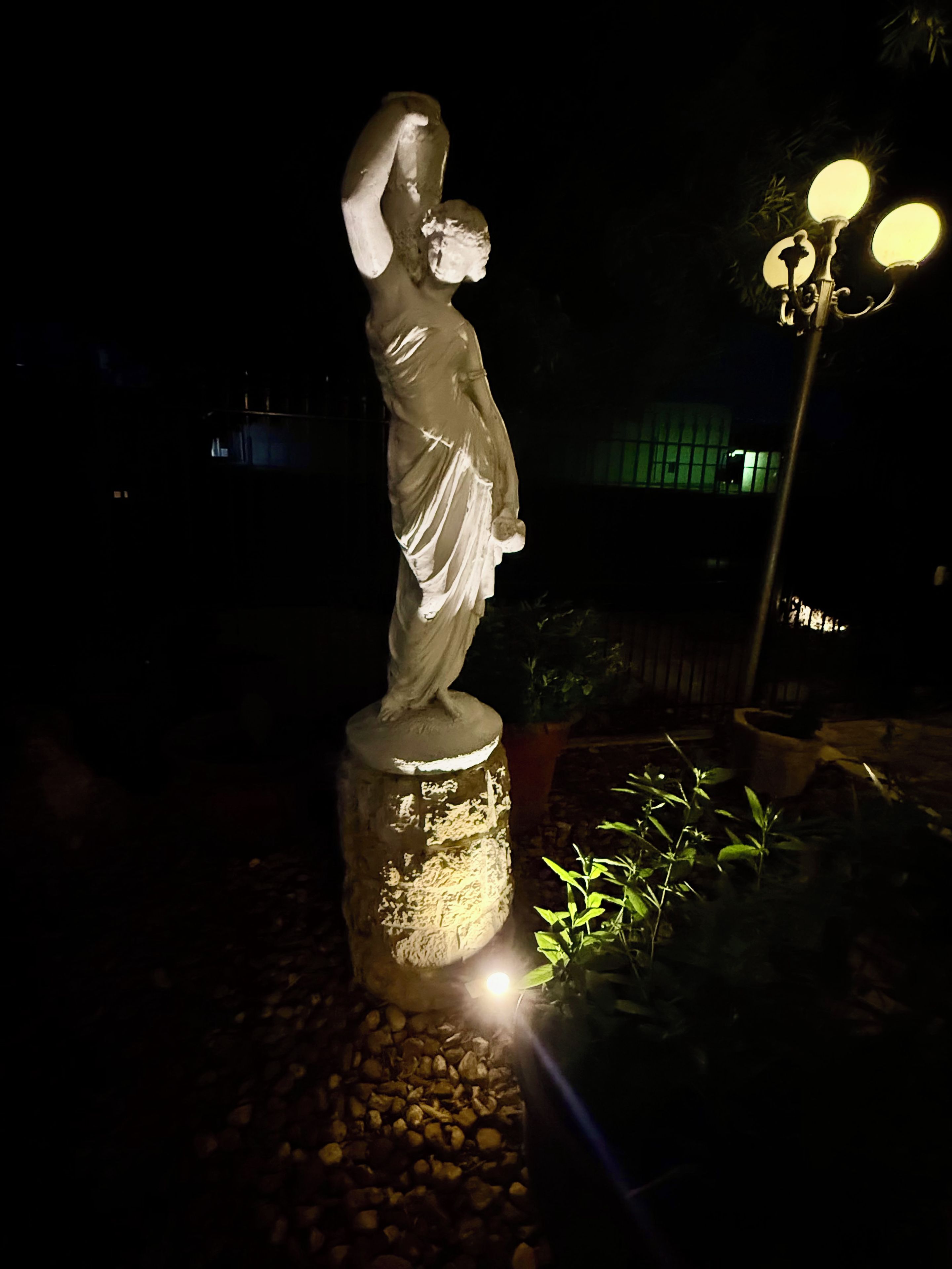 Eine Statue, die nachts beleuchtet wird, auf einem Steinsockel platziert, mit einem Laternenpfahl in der Nähe.