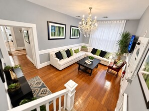 A modern living room with a white sofa, a coffee table, and a chandelier. There are framed pictures on the wall and a plant on the side. - Downtown Mansion 10 bedrooms/7.5 baths, 34+ people, 11car gated parking, Jacuzzi (San Antonio)