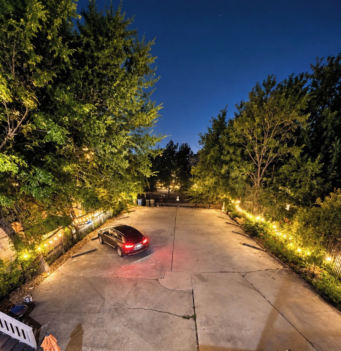 Un camino pavimentado que conduce a un acceso con puerta, rodeado de árboles e iluminado con luces colgantes.