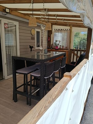 A wooden deck with a glass-top table and chairs, a hanging light fixture, and a view of the outdoors. - Mobile home 50 meters from the beach Natural surroundings Family vacations (Agde)