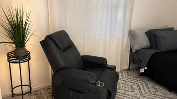Una sala de estar con una butaca reclinable negra, un tapete estampado, una mesita de noche con una planta y un sofá con almohadones.