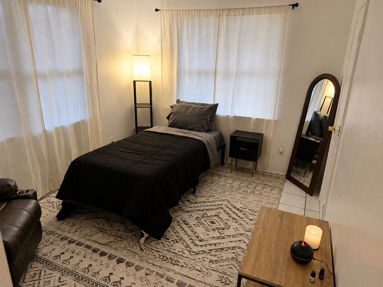 Un dormitorio con cama, mesita de noche, lámpara, espejo y una pequeña mesa de madera con una vela.