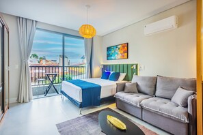 A modern hotel room with a balcony, a bed, a sofa, and a dining table. - Studio Saint Garden - Sunset - Pool with Ocean View - Dining (Florianópolis)
