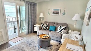 A living room with a sofa, ottoman, and a small table with a lamp. There is a view of the outdoors through a sliding door. - Oceanfront Condo Newly Renovated (Hampton)