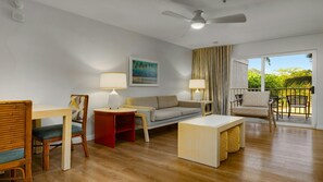 Una sala de estar moderna con sofá, sillas, una mesa de centro y una mesa de comedor. Hay un ventilador de techo, un cuadro en la pared y vistas a zonas verdes exteriores.