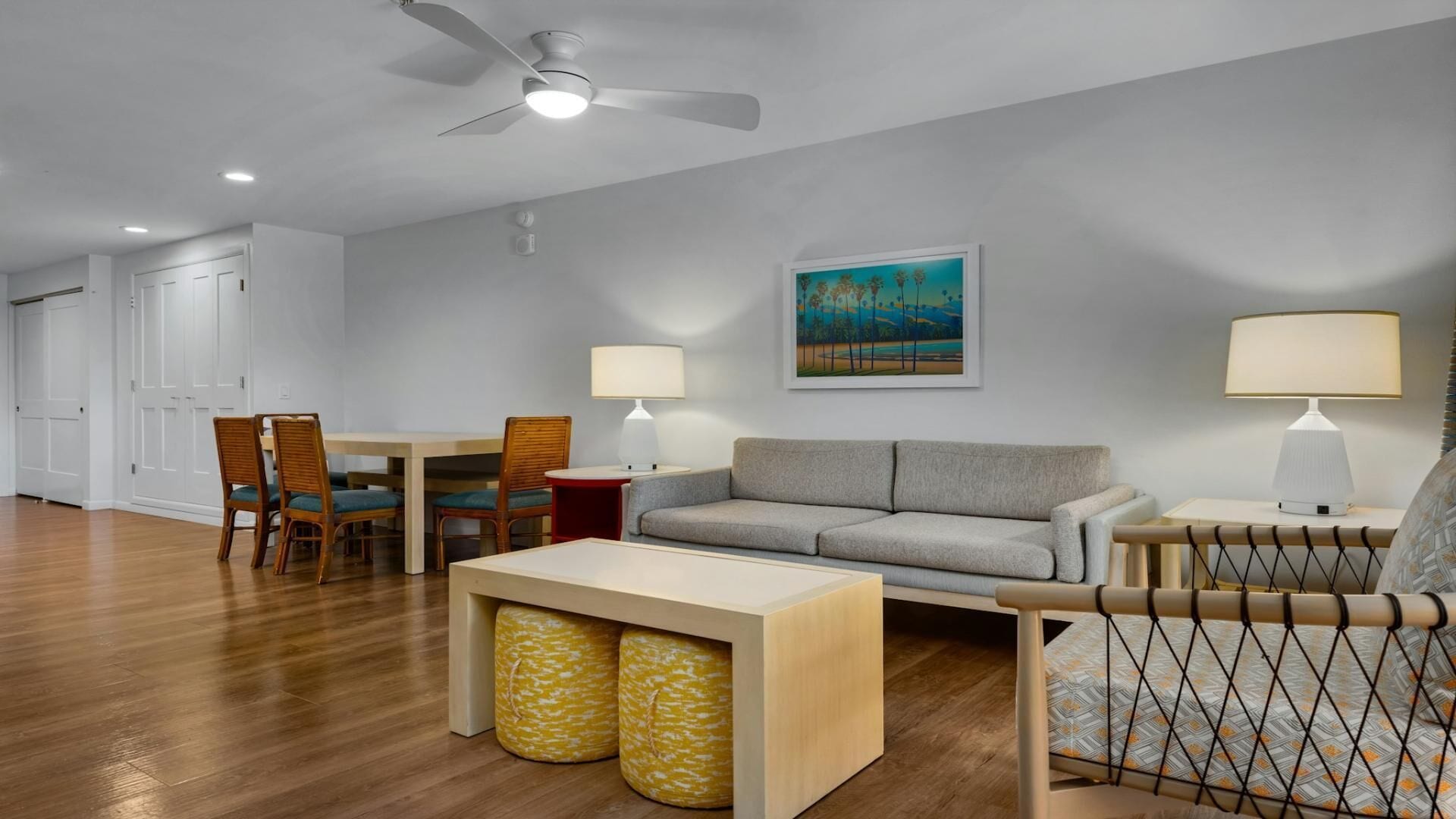 A modern living room with a grey sofa, wooden chairs, a dining table, and a ceiling fan.