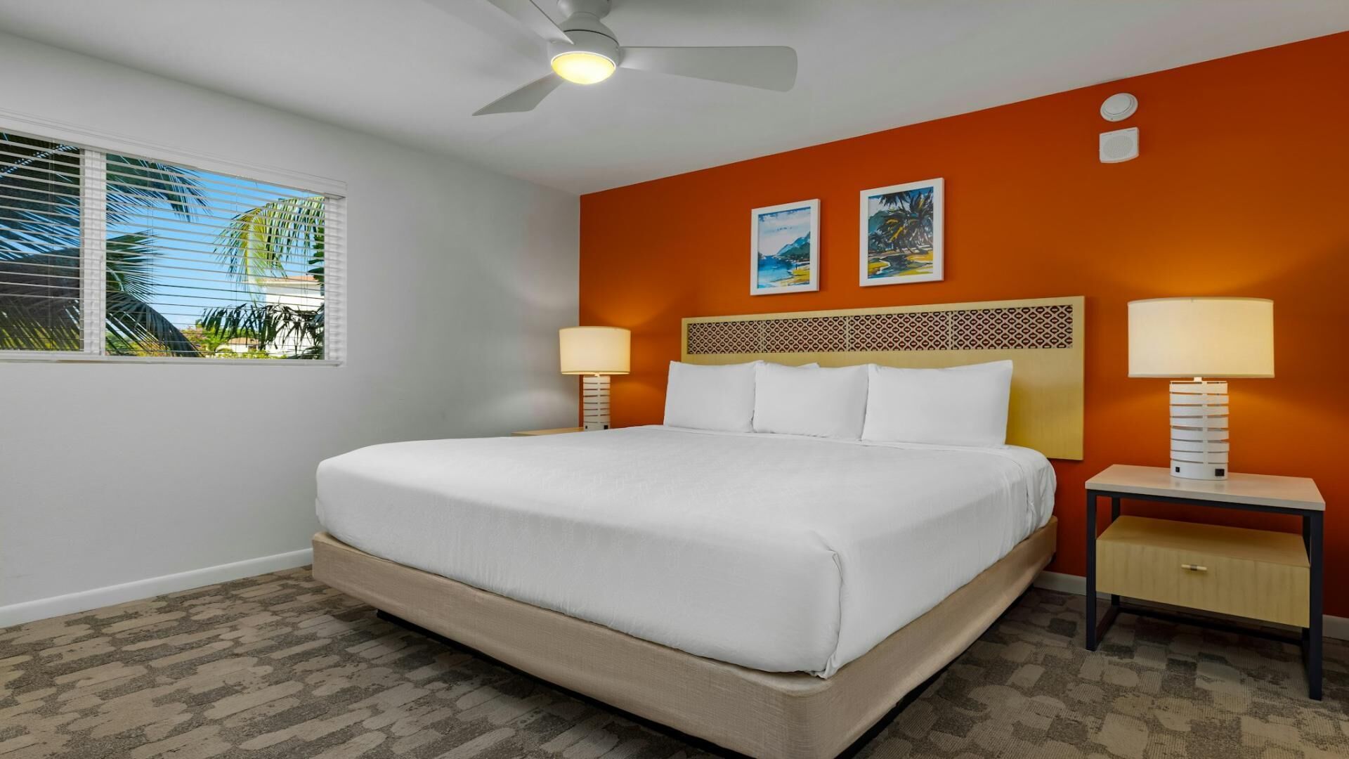 A hotel room with a large bed, two bedside tables, a ceiling fan, and two wall-mounted paintings.