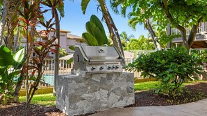 Un barbecue extérieur installé sur une base en pierre, entouré de plantes tropicales et d’une piscine.