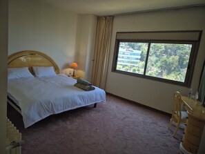 A hotel room with a bed, a desk, a lamp, a window with a view of buildings, and a carpeted floor. - Brilliant Beit meri  sea view  (Beit Mery)
