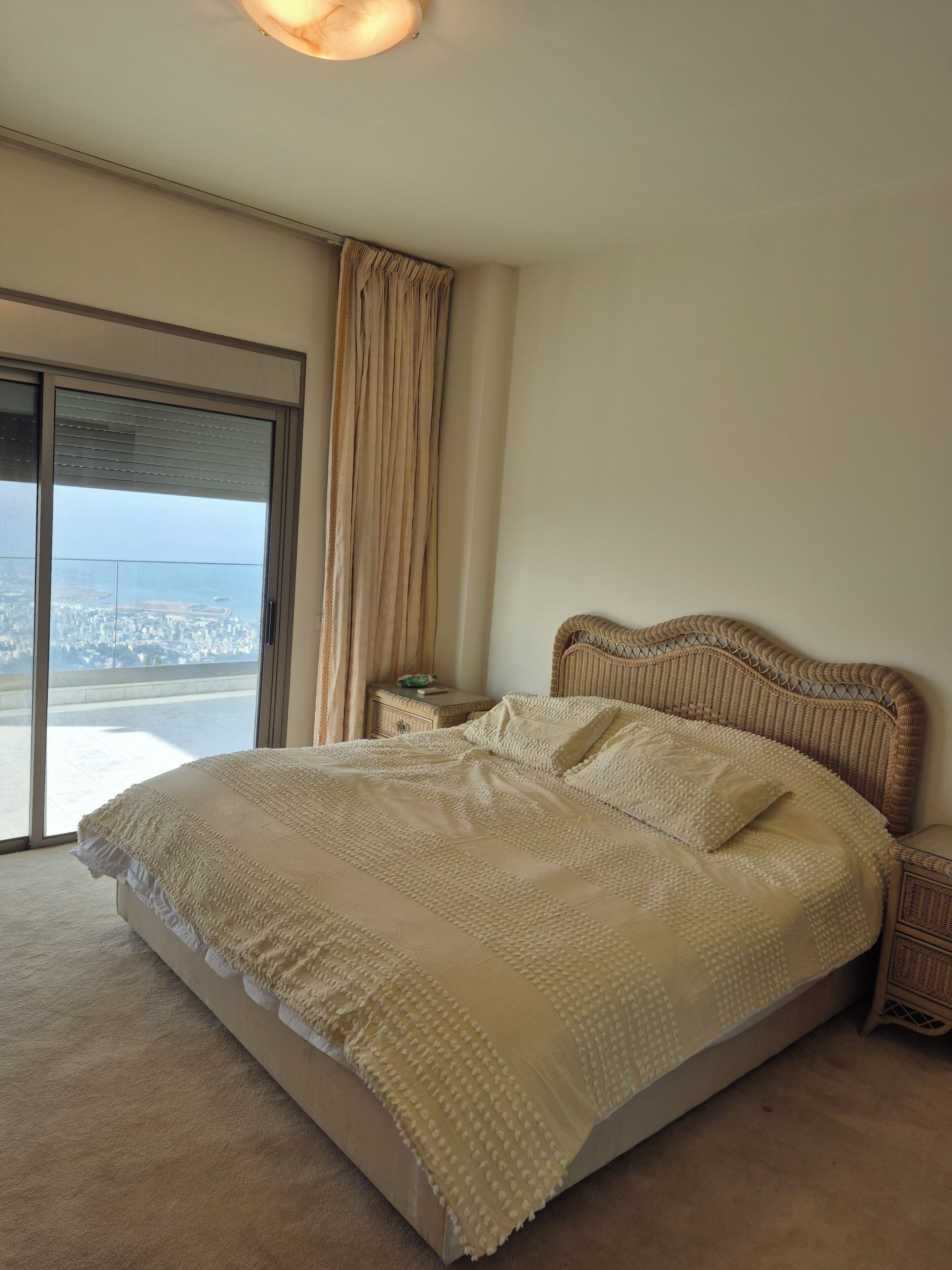 Un dormitorio con una cama grande, una mesita de noche, un cabecero de mimbre y vistas a través de un ventanal.