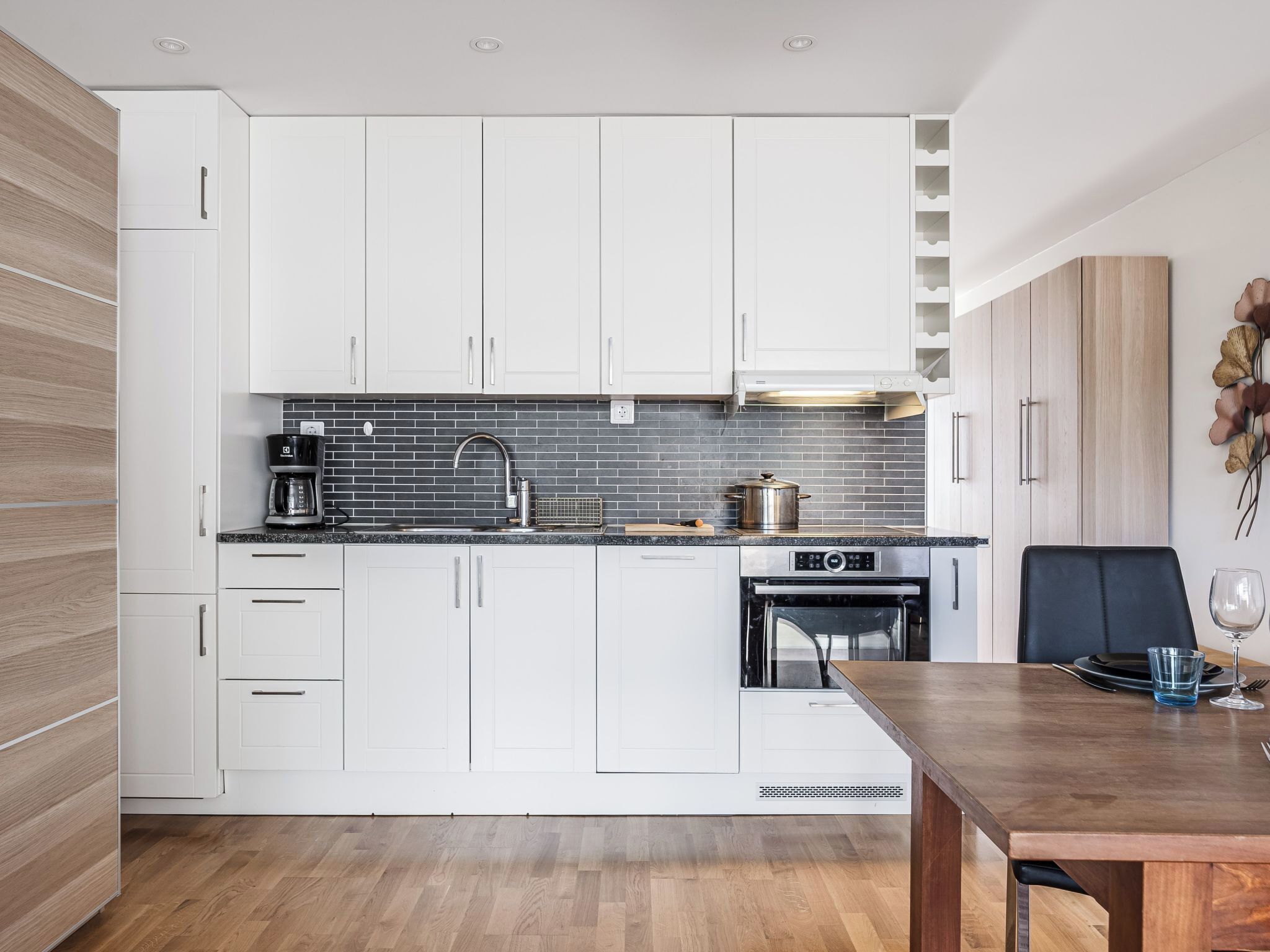 Een moderne keuken met witte kastjes, een zwarte achterwand en een houten eettafel.