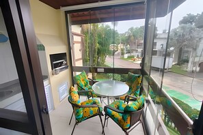 A balcony with a glass table and chairs, a TV, and a view of a residential area. - SHB Riviera Dreams com Piscina e Sauna (Bertioga)