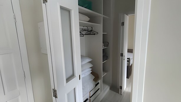 Un pasillo estrecho con una puerta blanca a la izquierda, que da a un armario con estantes y espacio para colgar.