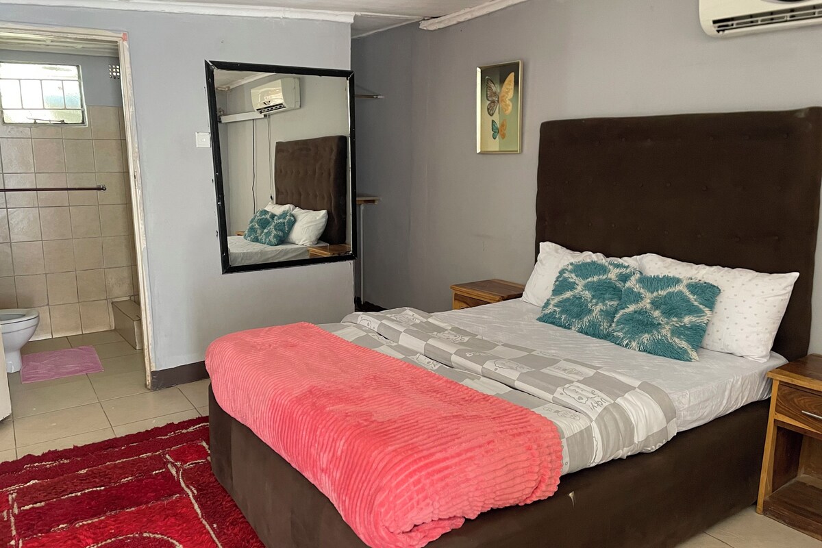 A bedroom with a bed, a nightstand, a mirror, a wall-mounted air conditioner, and a bathroom visible through an open door.