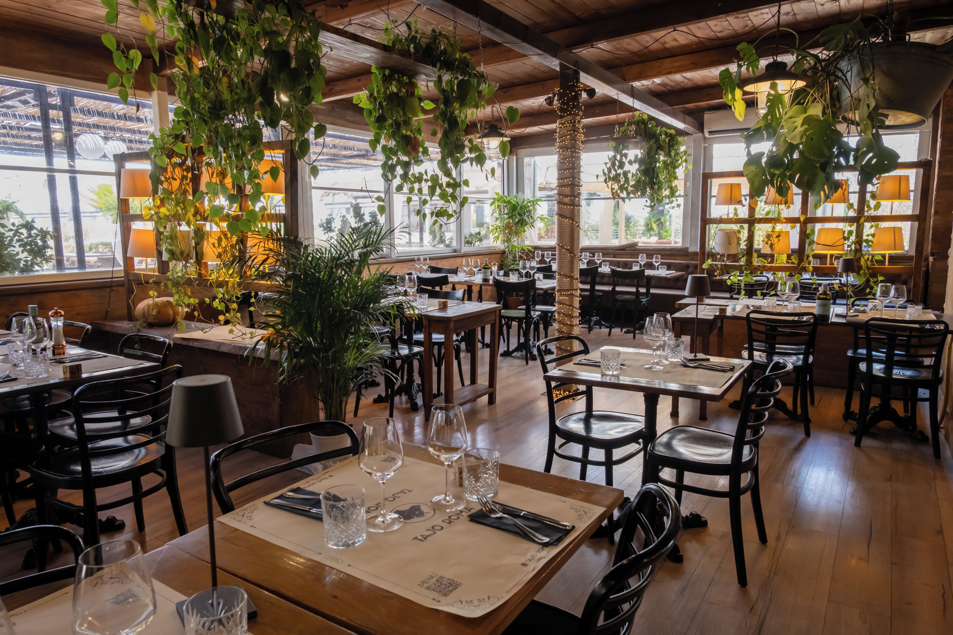 Un interior de restaurante con mesas y sillas de madera, plantas colgantes y vistas al exterior a través de amplios ventanales.