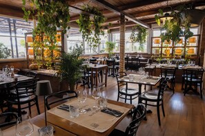 Un interior de restaurante con mesas y sillas de madera, plantas colgantes y vistas al exterior a través de amplios ventanales.