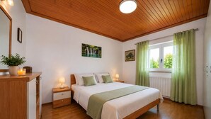 A bedroom with a wooden ceiling, a bed with white bedding, a nightstand with a lamp, a window with green curtains, and a plant on the dresser. - Gorgeous home in Selca with WiFi (Selca)