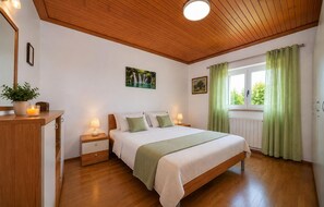 Ein Schlafzimmer mit Holzbalkendecke, einem Bett mit weißer Bettwäsche, einem Nachttisch mit Lampe, einem Fenster mit grünen Vorhängen und einer Pflanze auf der Kommode.