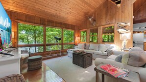 Interior de una cabaña de madera con ventanas grandes, un sofá, un reposapiés de mimbre y una lámpara.