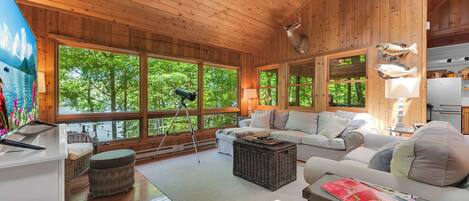 A wooden cabin interior with large windows, a sofa, a wicker ottoman, and a lamp.
