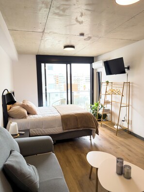 Una habitación de hotel moderna con cama, sofá, mesita de noche, televisión y balcón con planta.