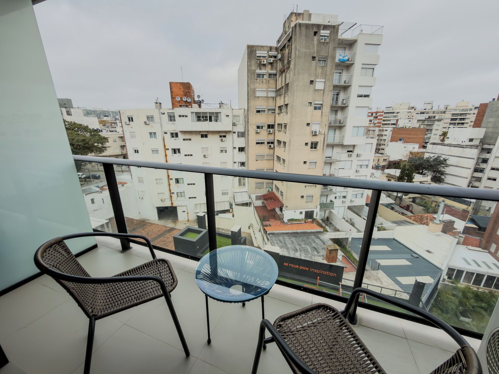 A balcony with two wicker chairs and a small table, overlooking a cityscape with multiple buildings.