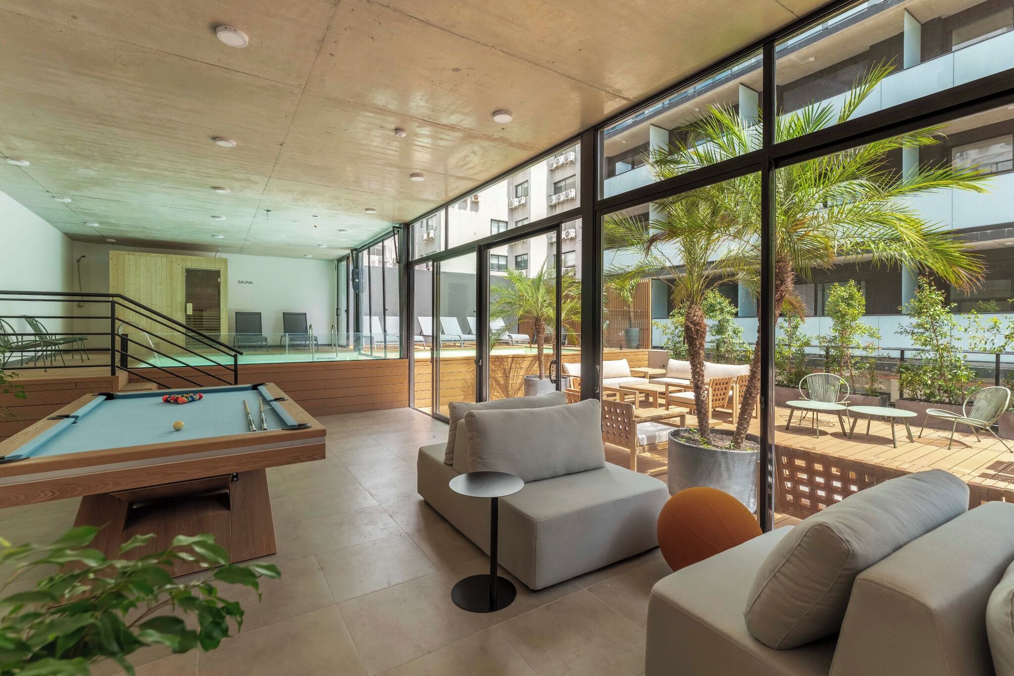 Un espacio interior moderno con mesa de billar, zona de estar y grandes ventanales con vistas a un patio.