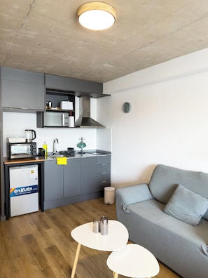 A compact kitchen with a microwave, kettle, and sink. There is a grey sofa and a small white table with two cups. - Modern WTC Studio Apartment | Gym + Pool (Montevideo)