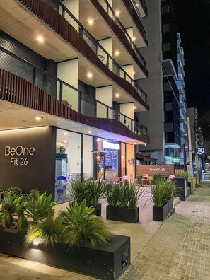 Un edificio moderno con un letrero que dice 'BeOne Fit 26', indicando un gimnasio. Hay zonas de estar al aire libre con plantas en macetas.
