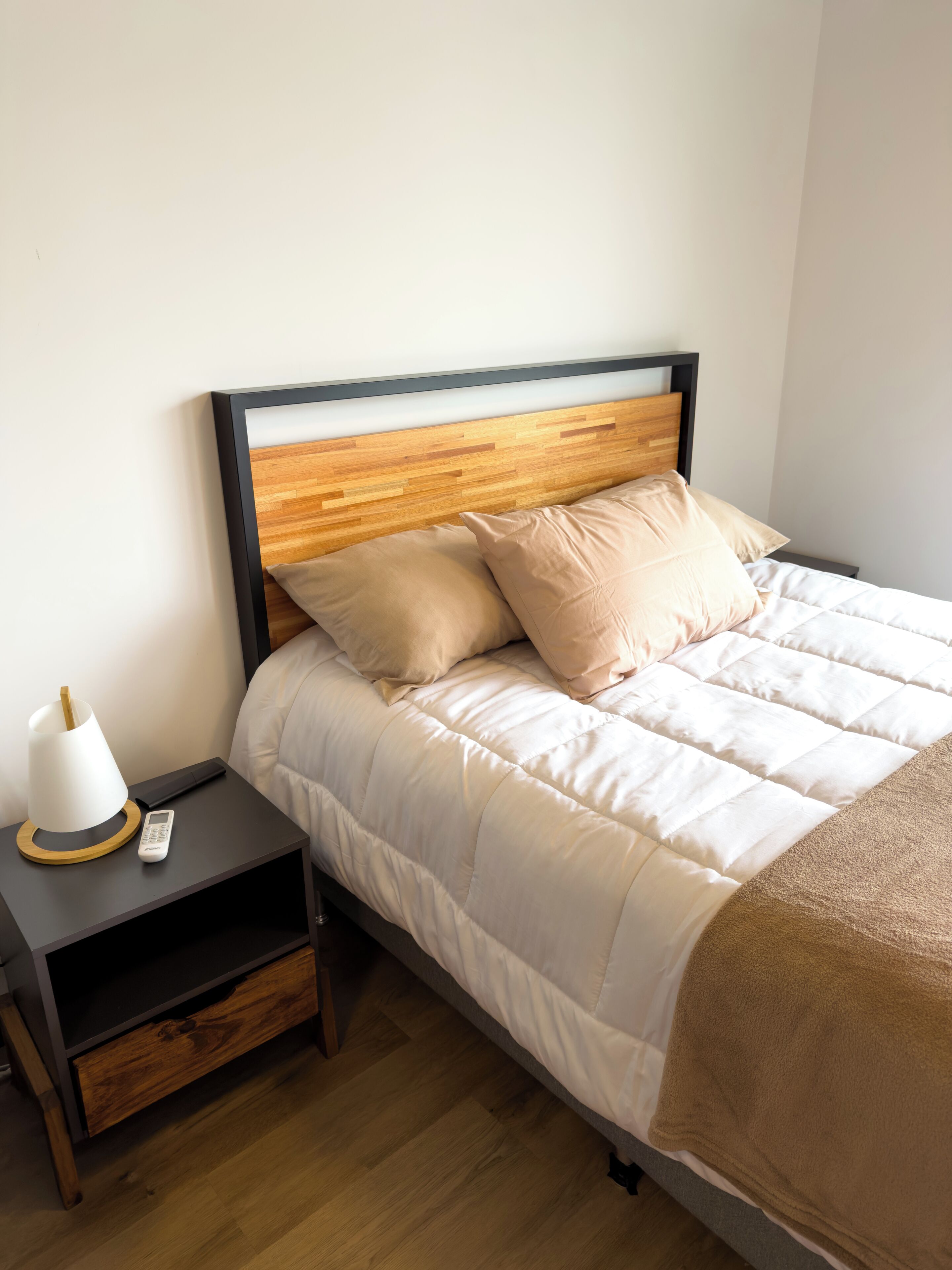 Uma cama bem arrumada com cabeceira de madeira, uma mesinha de cabeceira com abajur e um conjunto de gavetas.