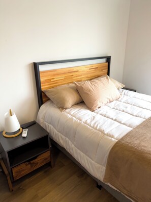 Una cama bien hecha con cabecera de madera, una mesita de noche con lámpara y una cómoda.