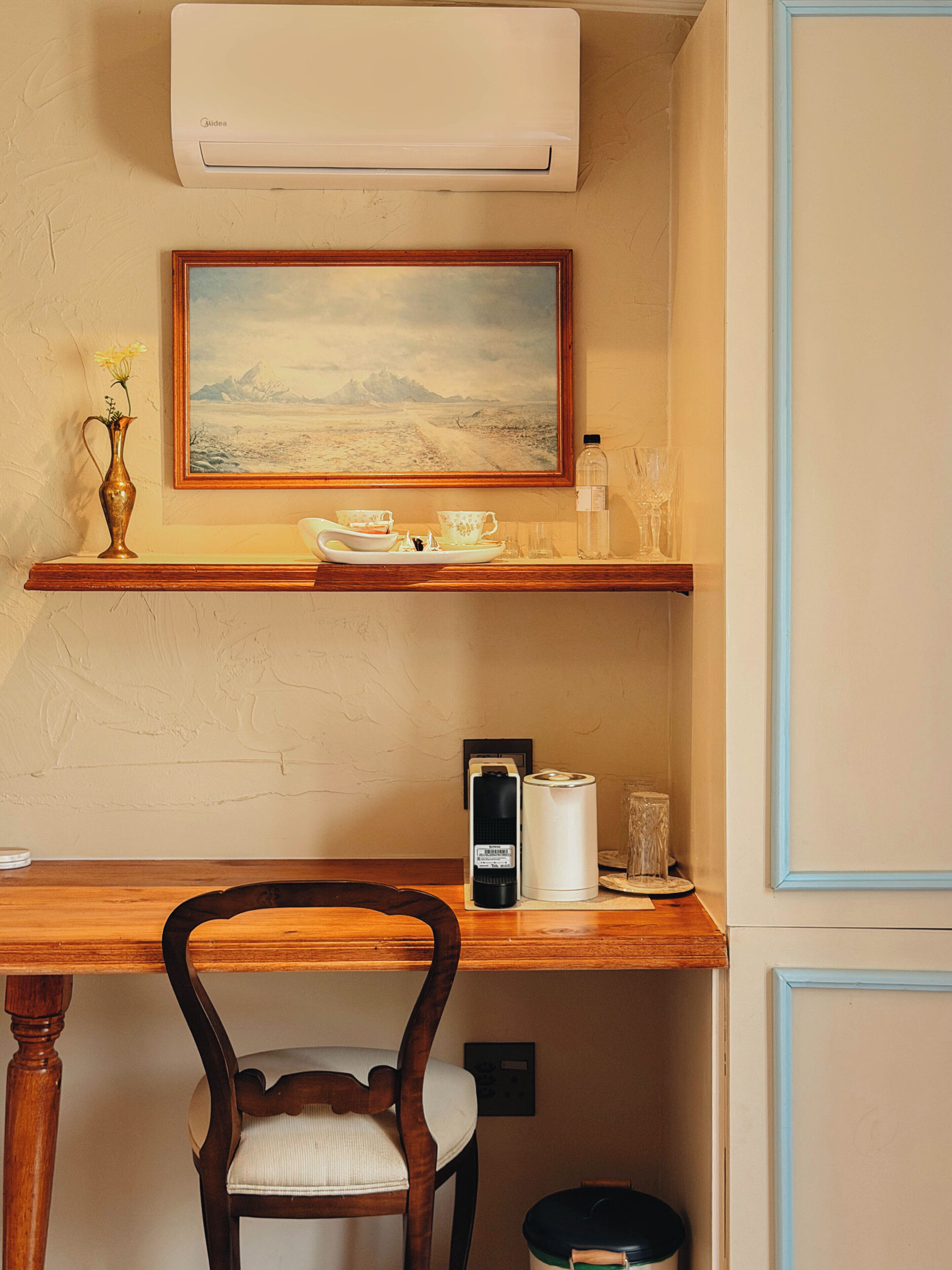 A room with a wooden desk, a chair, a framed painting, a vase with flowers, and a water bottle.