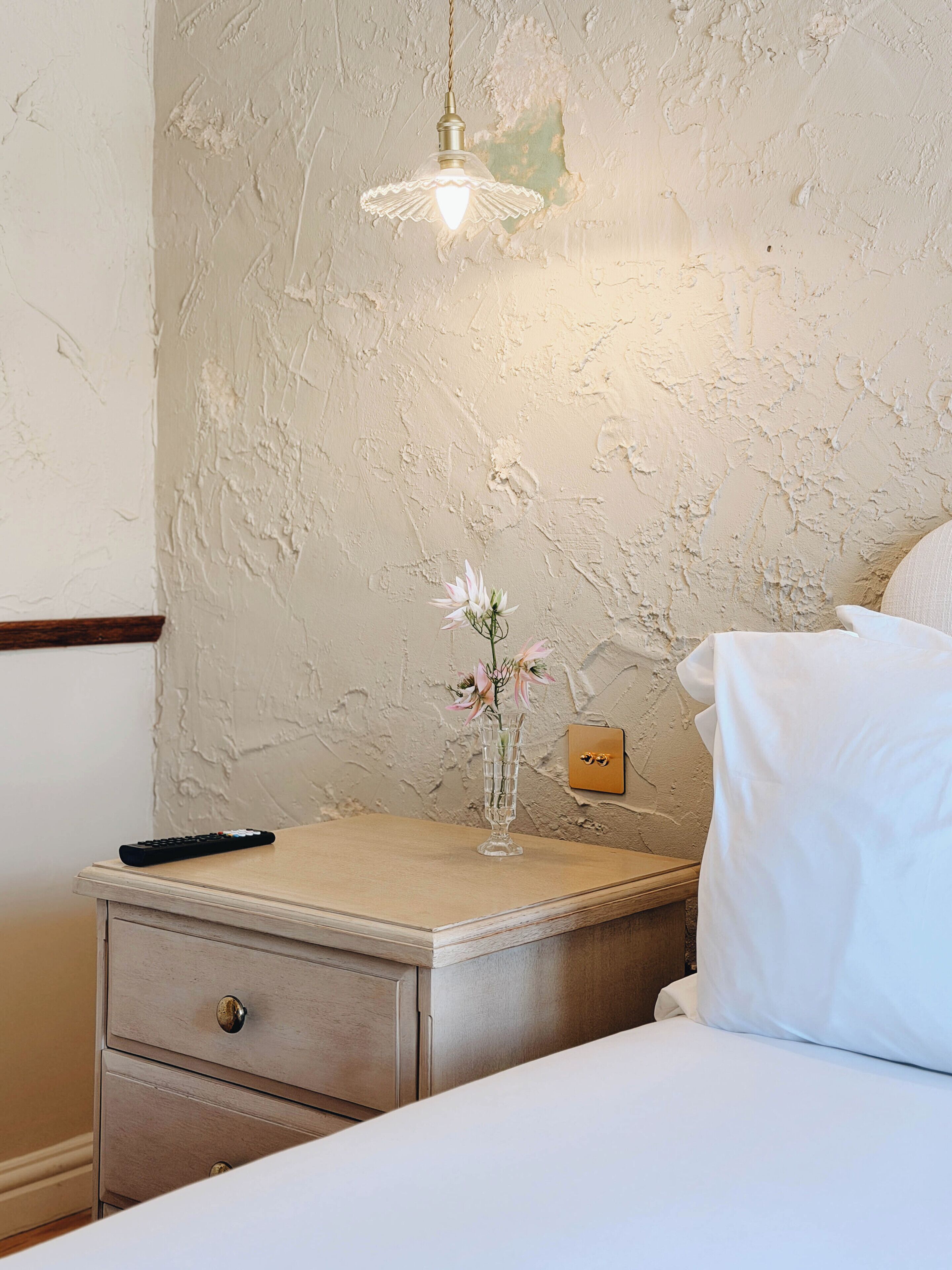 A bedroom with a bed, a nightstand, a vase with flowers, a remote control, and a wall socket.