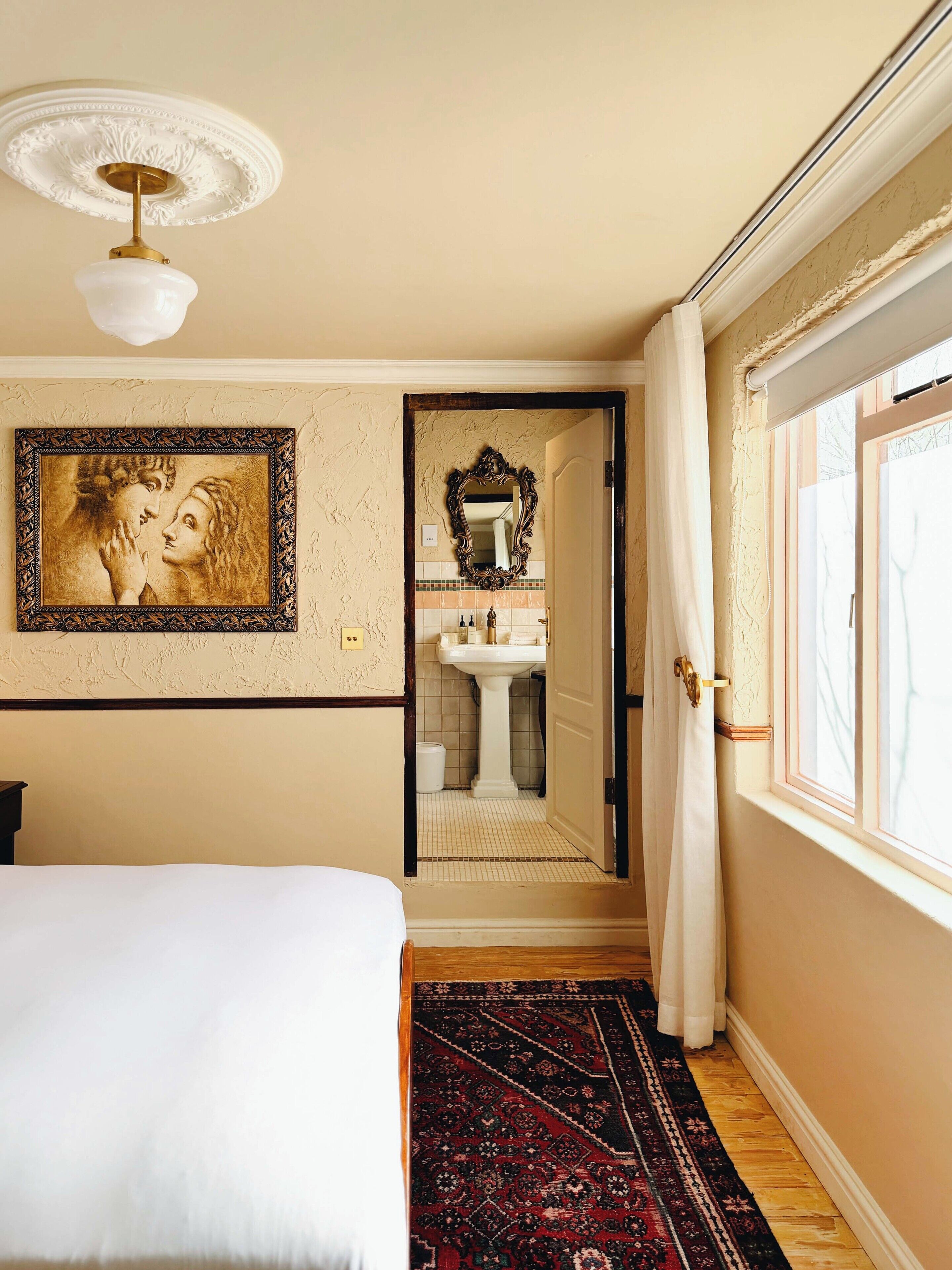 A bedroom with a bed, a large framed artwork, a bathroom with a mirror, and a window with curtains.