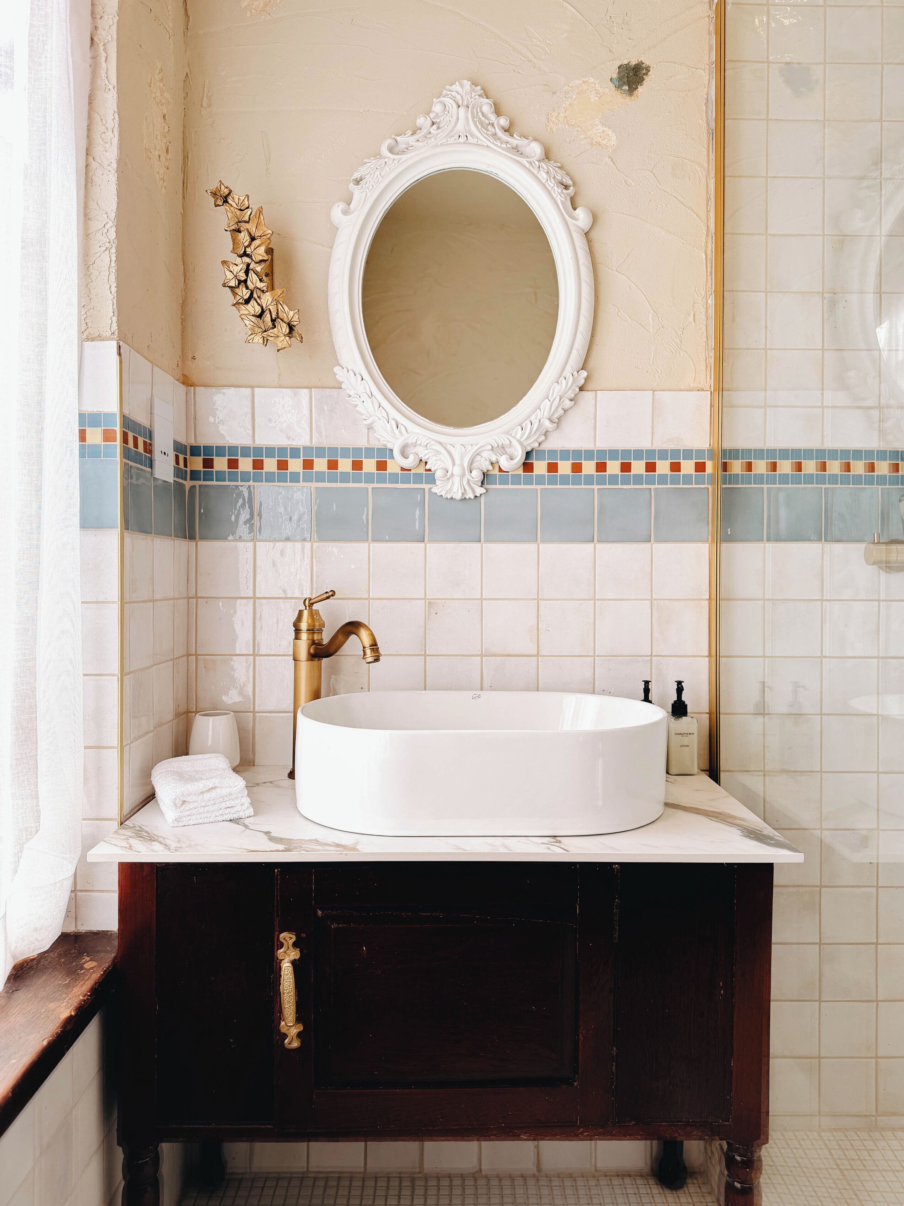A bathroom with a white sink, gold faucet, and a decorative mirror above it.