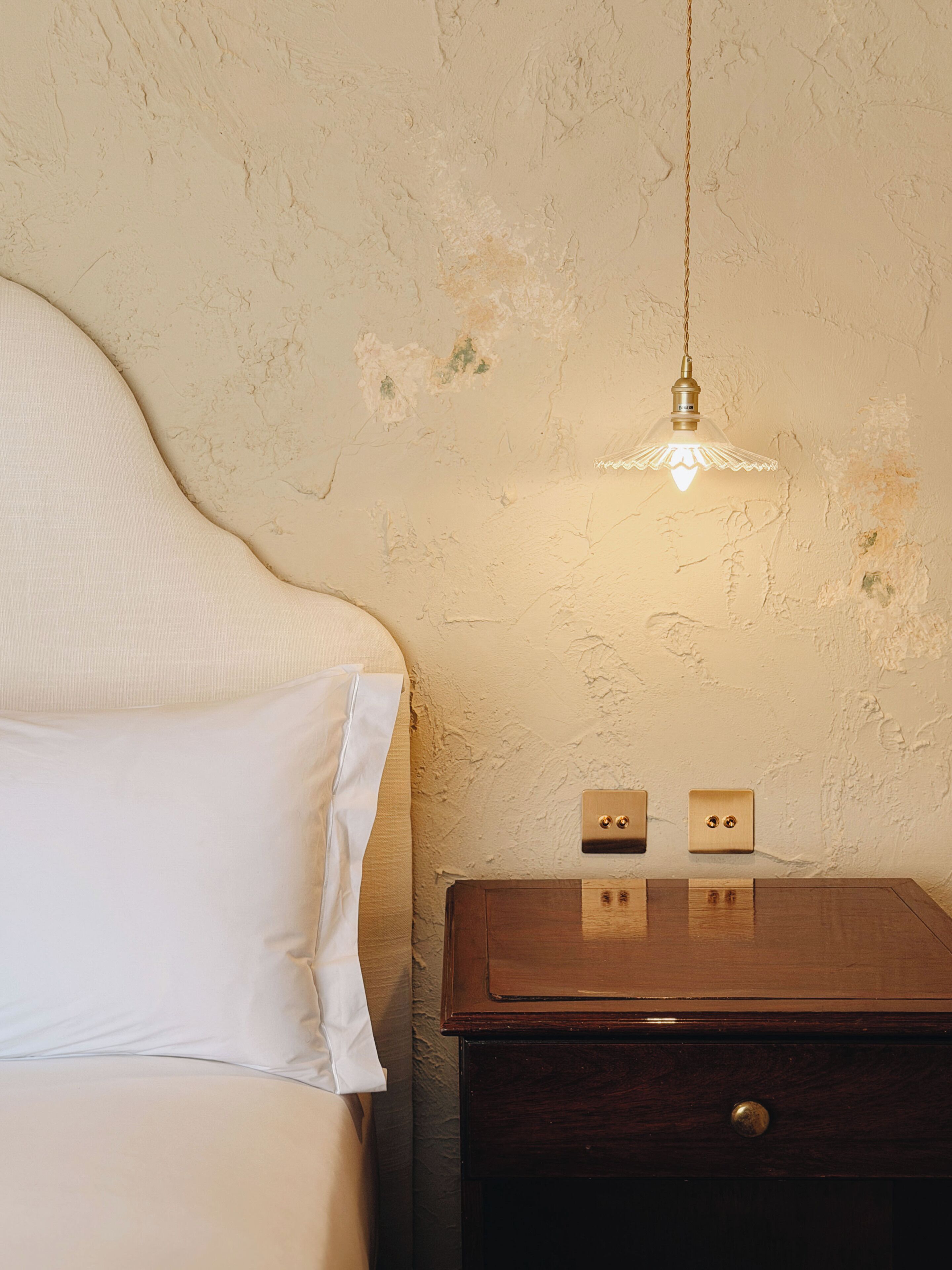 Uma cama com roupa de cama branca, uma mesa de cabeceira de madeira com dois interruptores de luz e um candeeiro suspenso.