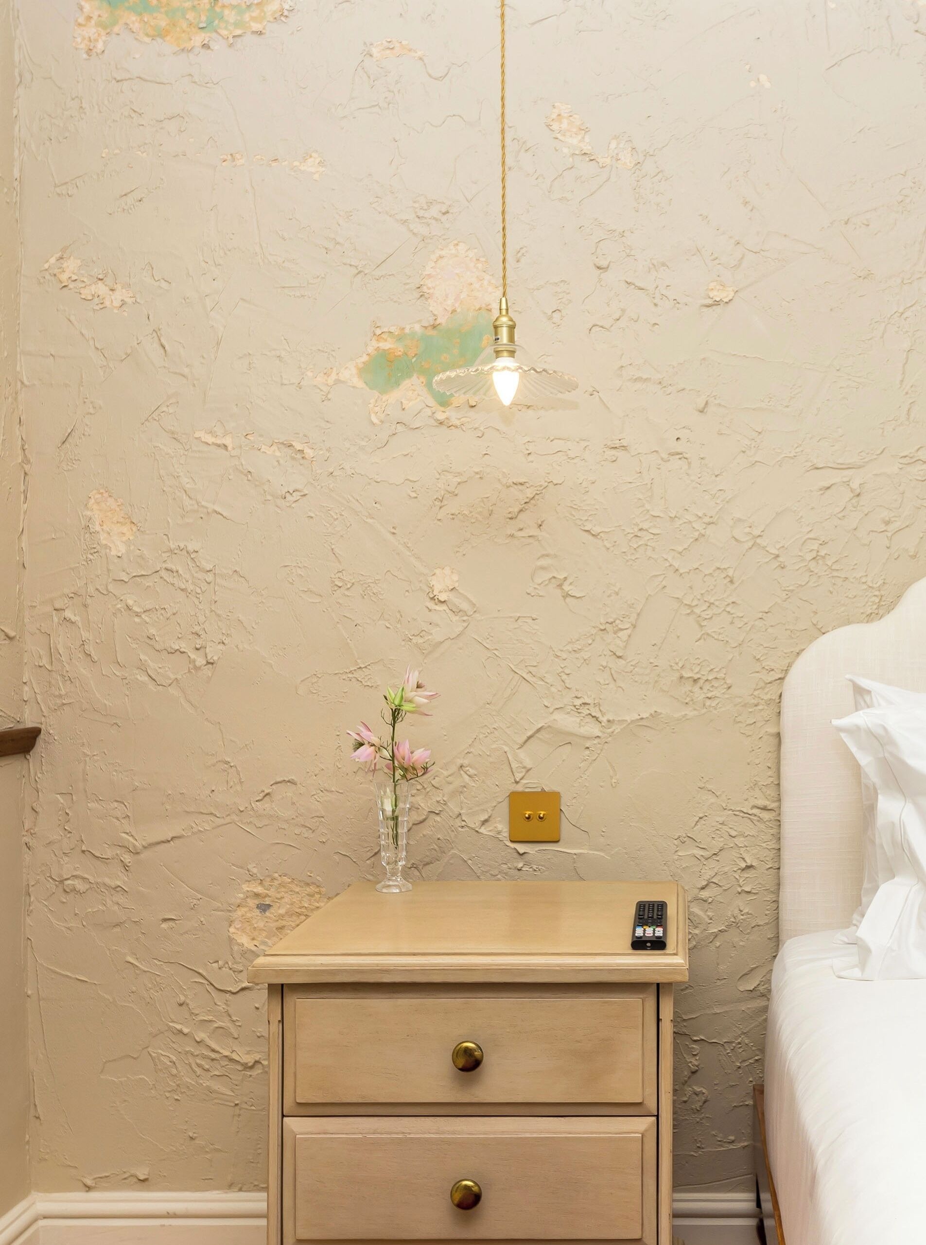 A bedroom with a bed, a bedside table with a vase of flowers, a lamp, and a wall with peeling paint.
