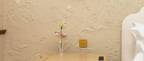 A bedroom with a bed, a bedside table with a vase of flowers, a lamp, and a wall with peeling paint.