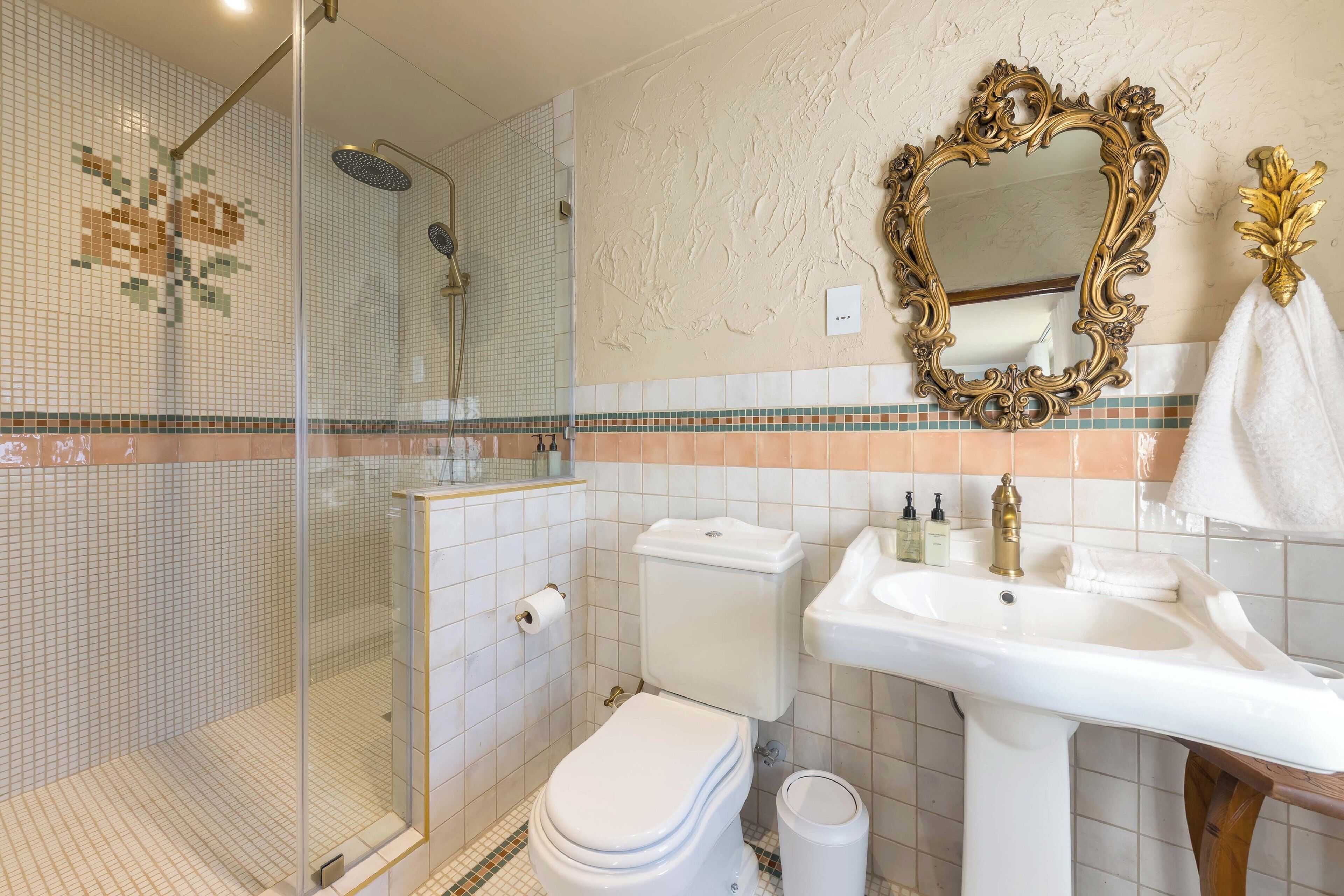Un bagno con doccia vetrata, un water, un lavandino con rubinetto dorato e uno specchio decorativo.